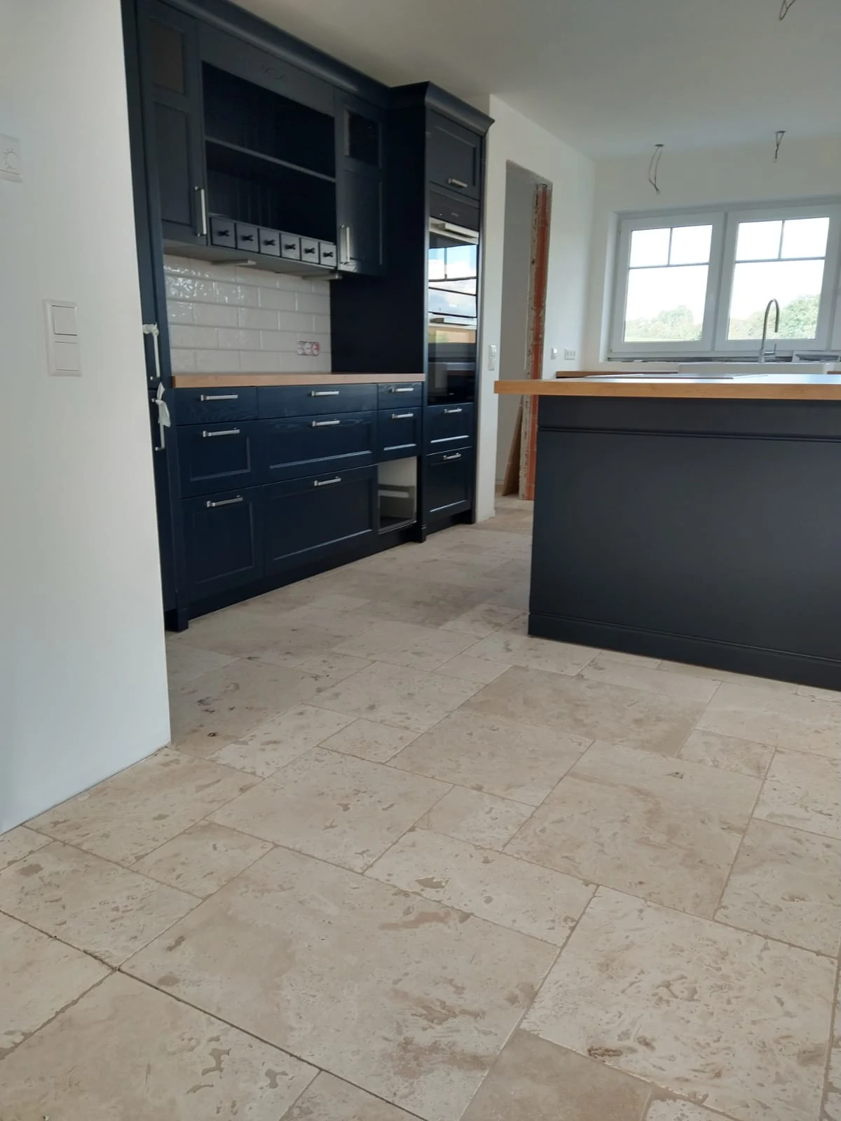 Dunkelblaue Küche mit Hochschränken, Kücheninsel und Spülbereich am Fenster, vom Eingang aus gesehen